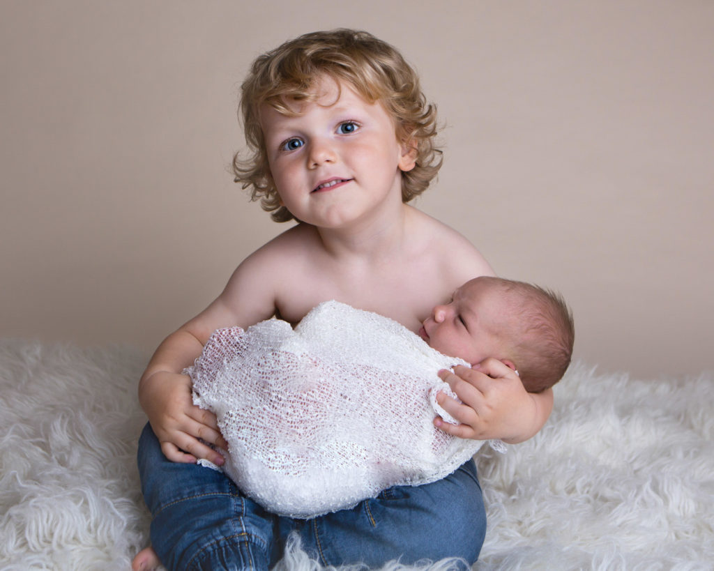 Big brother is holding his baby who is wrapped in a white wrap at their newborn photoshoot