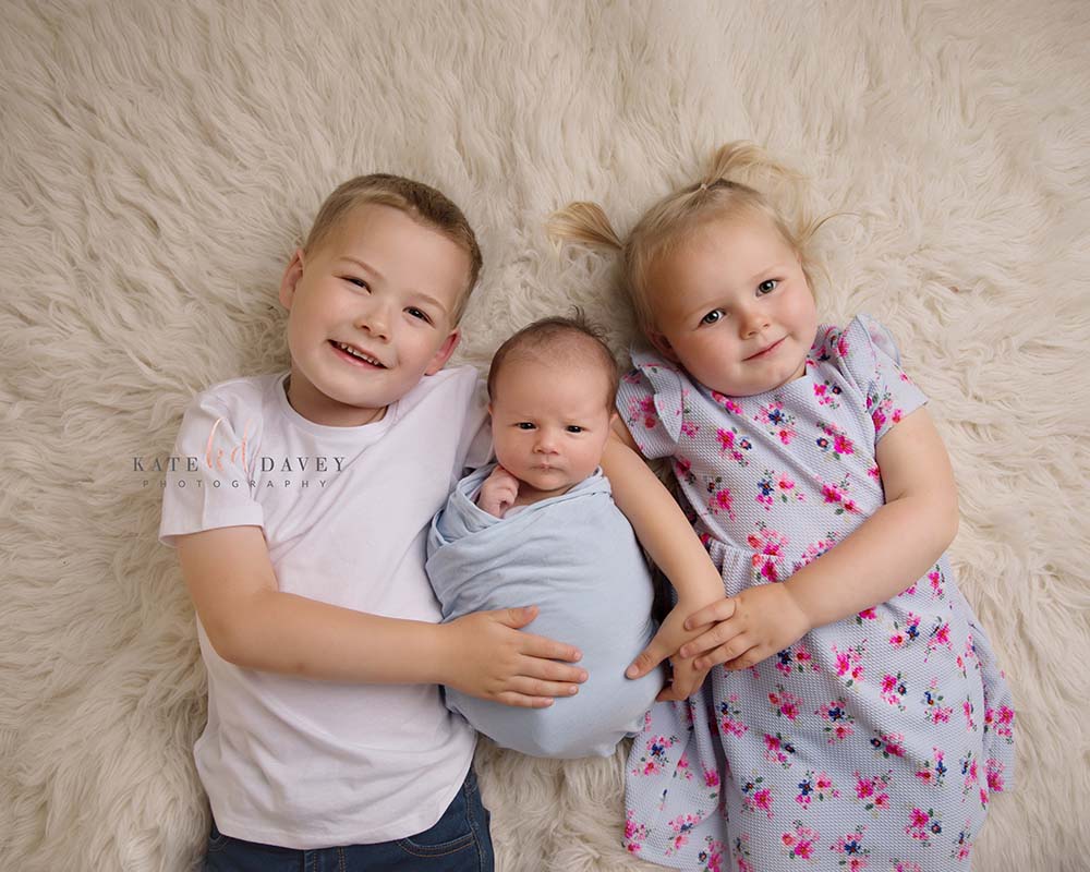 Three children lying down on a cream rub hugging their new baby brother